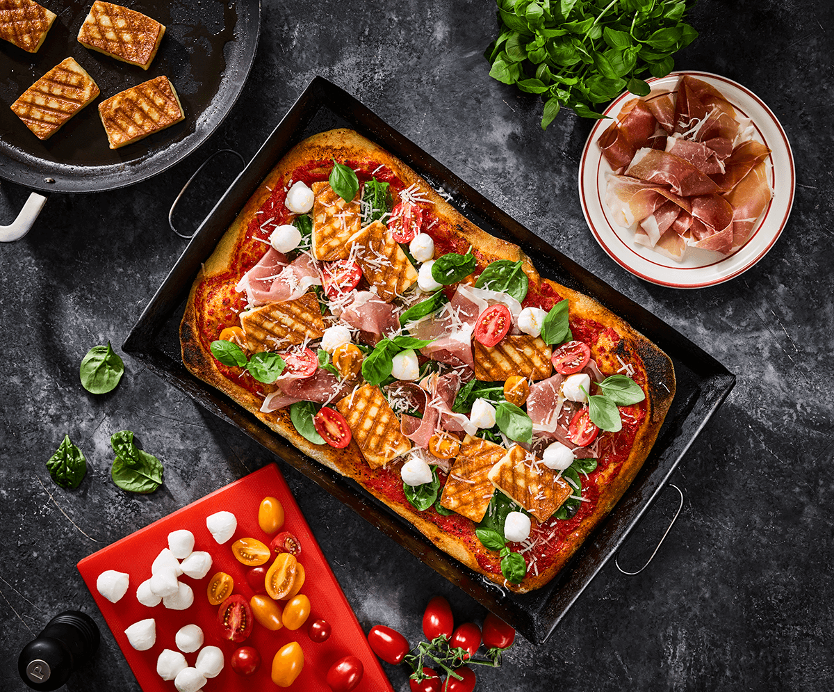 Fyrkantig gräddad pizza med toppings som ost, tomat, basilika och fat och skärbrädor med olika ingredienser runt om på en bänkskiva av mörk sten