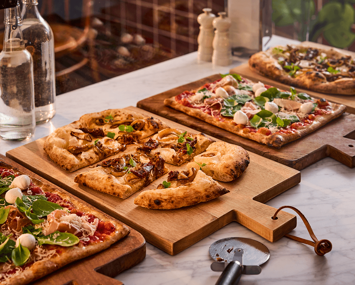 Gräddade pizzor i bitar på fyra skärbrädor av trä på en bänkskiva av sten och en pizzaslicer i förgrunden