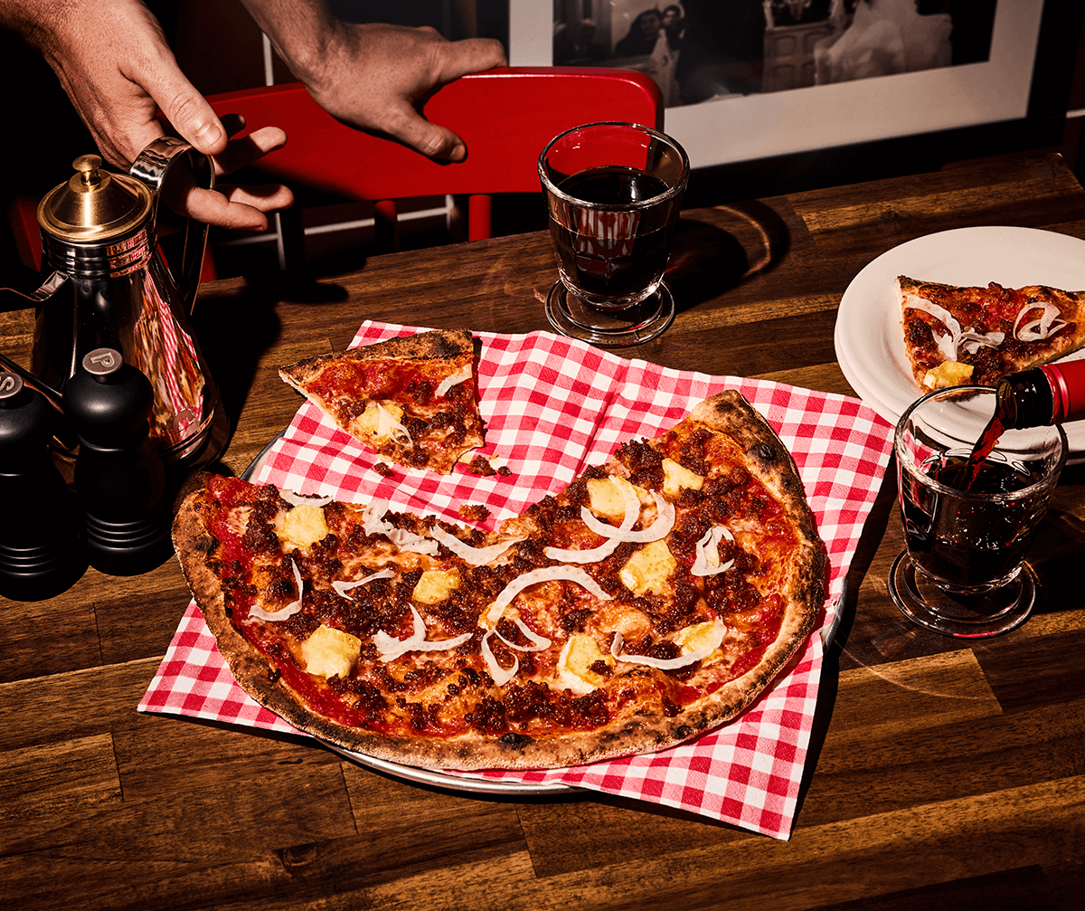 Gräddad pizza på rödvitrutig servett på ett träbord och en person bakom som är på väg att sätta sig ner