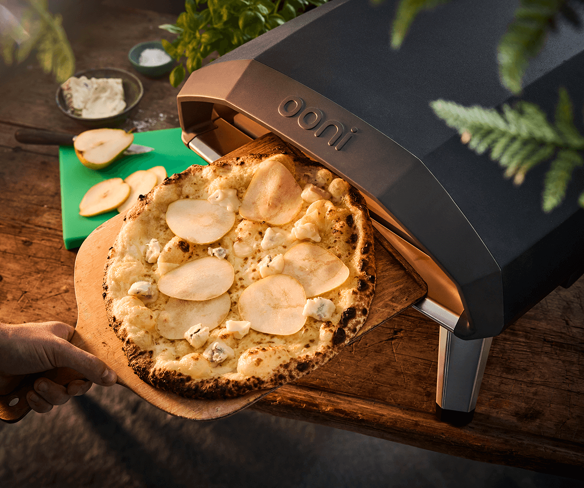 Gräddad pizza på väg ut ur en pizzaugn som står utomhus på ett bord