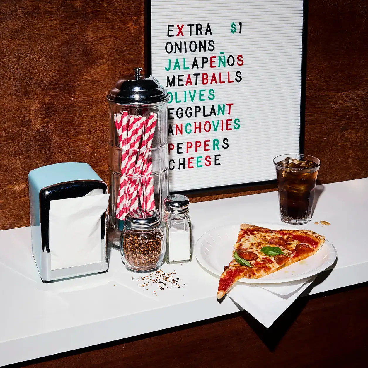 En pizzaslice på en tallrik på ett väggmonterat bord med servetthållare, sugrör, salt och peppar, ett glas läsk och en meny på väggen bakom