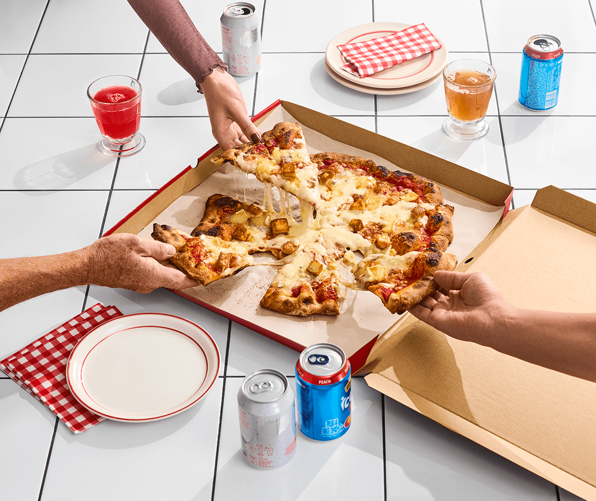 Tre händer som tar pizza slices från en pizzakartong på ett kaklat golv med tallrikar och rödvitrutiga servetter samt glas och läskburkar