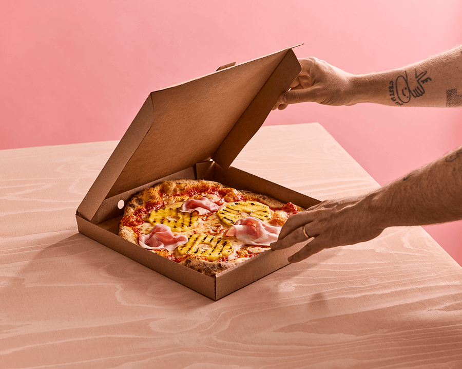 Två händer som öppnar en pizzakartong med en hawaiipizza i på ett bord med ljus duk
