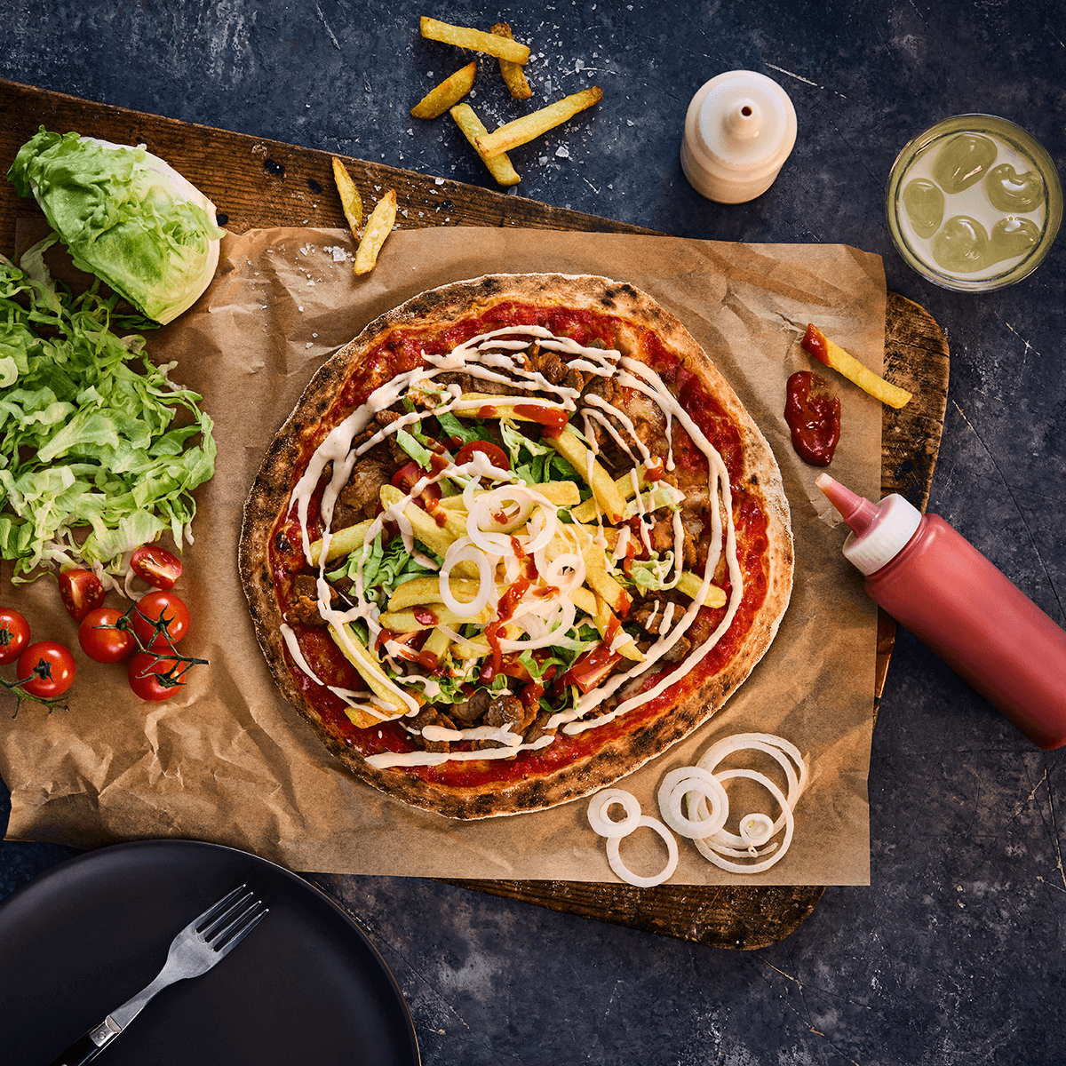 Gräddad kebabpizza på bakplåtspapper med sallad, tomat, pommes och kebabsås runt om på ett stenbord